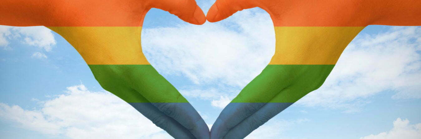 Hands painted as the rainbow flag forming a heart, symbolizing gay love on blue sky.